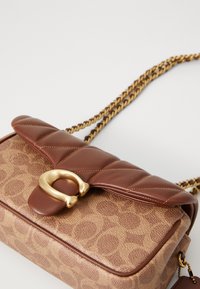 Brown quilted handbag with a gold circular clasp, featuring a textured pattern and a woven chain strap. Compact rectangular shape.