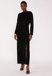 Robe longue en velours noir à manches longues avec un décolleté bateau et une fente sur le côté. Dotée d'une texture lisse et d'une silhouette ajustée. Portée avec des talons noirs.