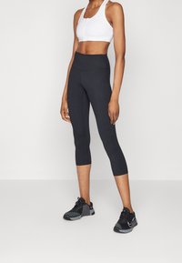 Schwarze Capri-Leggings mit hohem Bund und glatter Textur, kombiniert mit einem weißen Sport-BH mit Reißverschluss vorne und schwarzen Sportschuhen.