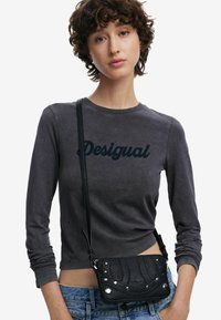 Schwarze Leder-Crossbody-Tasche mit strukturiertem Muster, silbernen Hardware-Akzenten und einem Reißverschluss, getragen über einem grauen Shirt mit "Desigual"-Text.