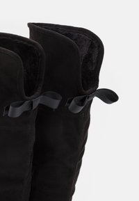 Stivaliin nabuk nero con rivestimento interno morbido e fiocchi decorativi in nera sul retro vicino ai bordi superiori.