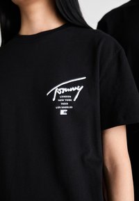 Musta T-paita, jossa on valkoinen "Tommy"-logo ja kaupunkien nimet Lontoo, New York, Pariisi, Los Angeles rinnassa, jonka päällä on henkilö, jolla on pitkät tummat hiukset.