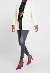 Vitt dubbelknäppt kavaj, grafisk svart t-shirt, grå smala jeans med paneldetaljer och lila mocka stiletthälar.