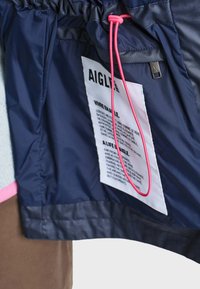 Intérieur d’une veste bleu marine avec une étiquette rectangulaire blanche portant la mention AIGLE et un texte, avec un cordon réglable rose et une tirette de fermeture éclair en métal.