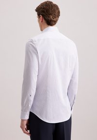 Man in een witte overhemd met lange mouwen en dunne verticale strepen, gezien vanaf de achterkant, gecombineerd met donkere broek tegen een effen achtergrond.