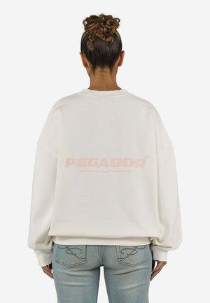 Kvinde med krøllet hår sat op i en knold, iført en løs hvid sweatshirt og lyseblå jeans, vist bagfra mod en ensfarvet baggrund.