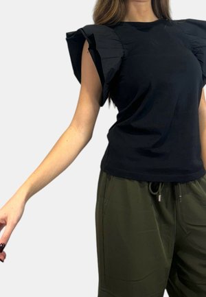 Kobieta ubrana w czarny top z dużymi falbanami na rękawach oraz oliwkowozielone spodnie z troczkiem, stojąca na tle jednolitego jasnego tła.