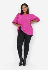Fuchsia Bluse mit kurzen puffärmel, einem V-Ausschnitt, kombiniert mit schwarzen engen Hosen und schwarzen Ankle-Boots, vor einem weißen Hintergrund.
