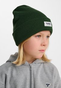 Grön stickad mössa med vikbar kant och en framträdande vit logotagg. Ämnet bär en ljusgrå zip-up hoodie.