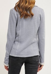Grå texturerad jacka med ett zigzagmönster, figursydd design och långa ärmar. Har en urklippt krage och kontrasterande manschetter.