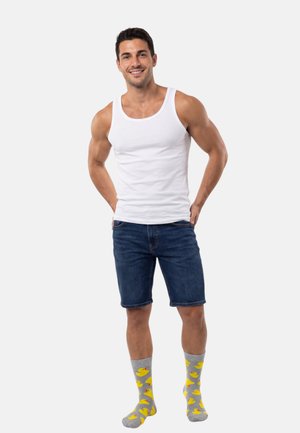 Valkoinen hihaton toppi, farkkushortsit ja harmaat sukat keltaisilla ankka-kuvioilla. Malli seisoo käsivarret lanteilla ja hymyilee neutraalia taustaa vasten.