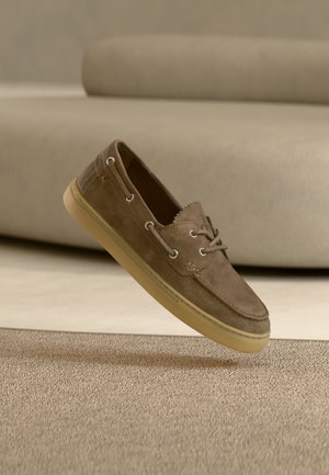 Chaussure bateau en daim marron avec semelle en caoutchouc beige flottant au-dessus d'un tapis beige texturé, devant un canapé beige assorti.