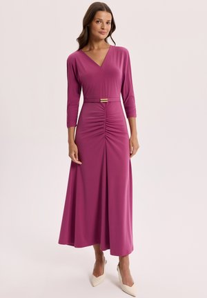 Kobieta ubrana w magentową sukienkę z marszczeniami na długich rękawach, z dekoltem w serek i paskiem, w połączeniu z białymi szpilkami o spiczastym nosku na jednolitym tle.