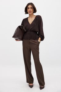 Blusa marrone plissettata con scollatura a V profonda e vita sottile, abbinata a pantaloni marroni su misura e una borsa coordinata. Le scarpe con tacco completano l'outfit.