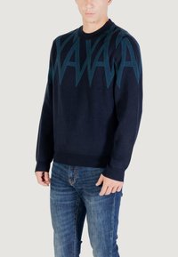 Homme portant un pull bleu marine avec un motif en zigzag bleu sur la poitrine et les manches, associé à un jean bleu, se tenant devant un arrière-plan uni.