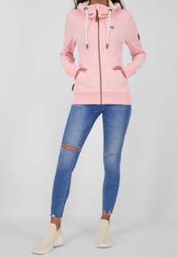 Růžová mikina na zip s vysokým límcem, šňůrkami a logem; kombinována s modrými roztrhanými skinny džíny a béžovými nazouvacími teniskami.