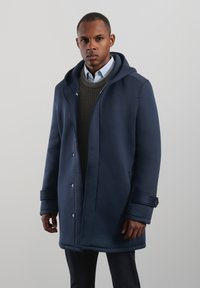 Navy blue hooded coat in smooth fabric with a relaxed fit, featuring a zip front, button cuffs, and a textured olive green sweater underneath.