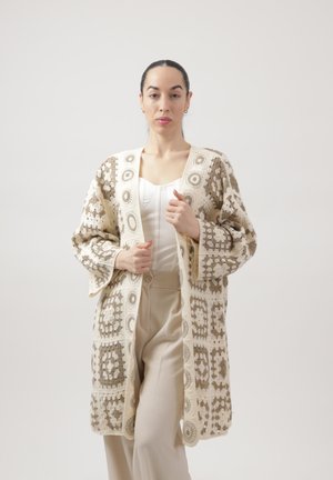 Donna che indossa un cardigan beige a crochet con motivo sopra una maglia bianca e pantaloni beige, in piedi contro uno sfondo uniforme.