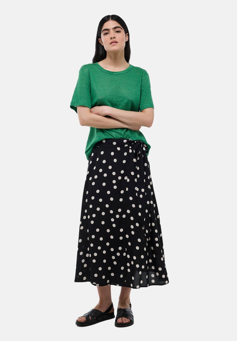 T-shirt texturé vert à manches courtes associé à une jupe portefeuille noire à pois avec un détail noué et des sandales noires à enfiler.