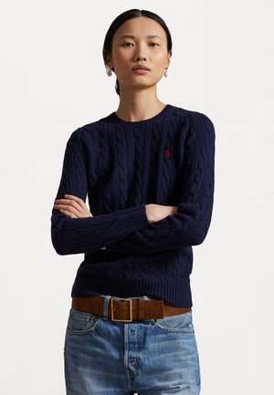 Pull en maille torsadée bleu marine avec un col rond et des manches longues, arborant un petit logo rouge sur la poitrine. Porté avec une ceinture marron et un jean.
