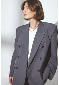 Blazer grigio doppiopetto con grandi bottoni neri, revers affilati e spalle strutturate. Indossato sopra un top bianco, con una finitura in tessuto liscia.