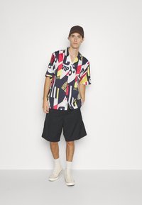 Kortærmet skjorte med et dristigt grafikmønster i rød, gul og sort; parret med sorte shorts og hvide sneakers.