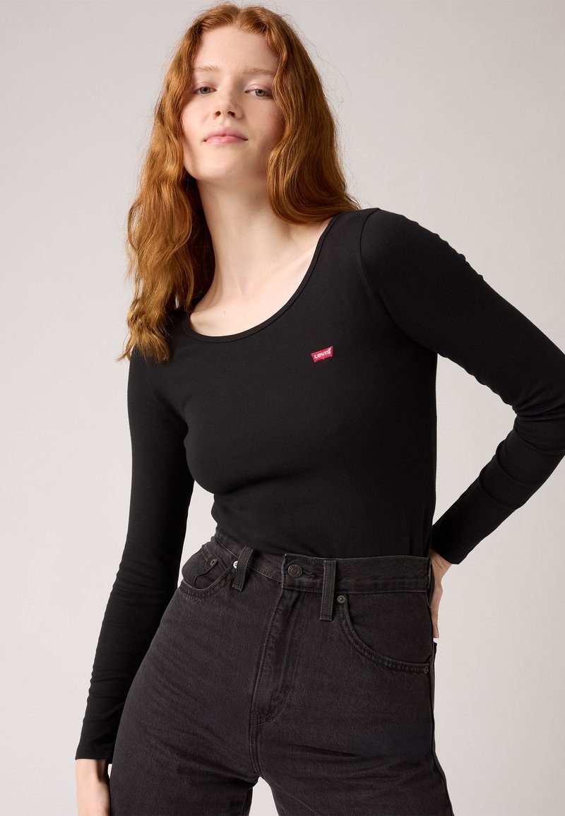 Svart langermet skjorte med scoop-hals og en liten rød Levi's-logo, kombinert med høytlivs svarte jeans i teksturert stoff.