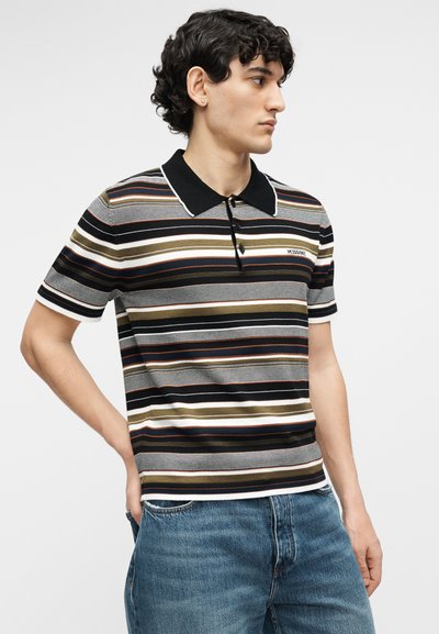 Νεαρός άνδρας που φοράει κοντομάνικο ριγέ πόλο με μαύρο γιακά, στέκεται με το ένα χέρι στην τσέπη, στραμμένος προς τα δεξιά.