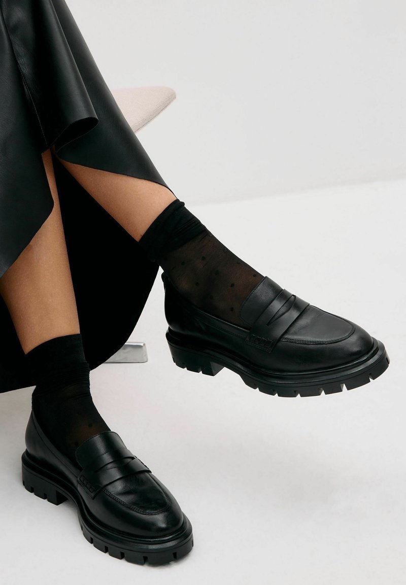 Personne portant des mocassins en cuir noir épais et des chaussettes noires transparentes à pois, assise les jambes croisées sur un fond clair.