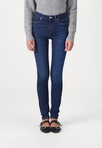 Pantalones vaqueros ajustados de mezclilla azul con cintura alta, que cuentan con cinco bolsillos, costuras tonales y cierre frontal con botón. Se llevan con un suéter gris y unos bailarinas negras.