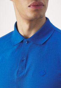 Polo bleu en tissu texturé, avec un col boutonné, des boutons contrastants et un petit emblème sur la poitrine.