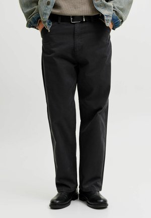 Homme portant un pantalon droit noir, ceinture noire, chaussures noires, chemise grise et veste en jean bleue avec les mains dans les poches.