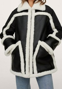 Sort læderjakke med hvid pelsforing, der har en lynlås foran, store lommer og en bred krave. Struktureret og oversized pasform.