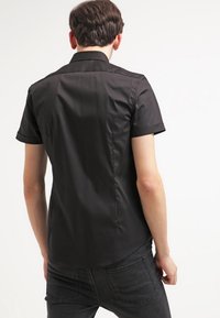 Czarna koszula z krótkim rękawem, wykonana z gładkiego materiału, z klasycznym kołnierzykiem i złożonymi mankietami, pokazana z tyłu. Brak widocznych wzorów i elementów metalowych.