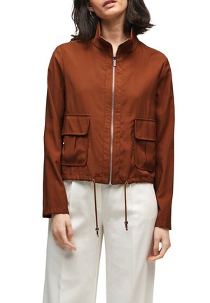 Kvinde iført en brun zip-up jakke med to forlommer med klap og snore i kanten, parret med hvide bukser med vide ben.