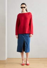 Červený vlnený sveter s boxovým strihom, spárovaný s modrou džínsovou midi sukňou s predným rozparkom. Outfit dopĺňajú červené sandále na podpätku.