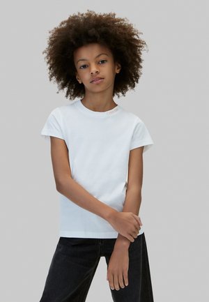 Enfant aux cheveux bouclés portant un t-shirt blanc à manches courtes et un pantalon noir, debout devant un fond gris uni.
