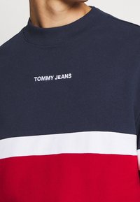 Osoba v námořnickém, bílém a červeném barevném bloku mikiny se vyšitým nápisem "TOMMY JEANS" na hrudi.