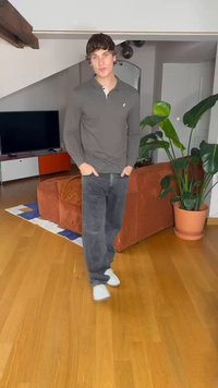 Srajca z dolgimi rokavi temno sive barve, črne jeans hlače, bele teniske. Notranje okolje z lesenim tlakom, oranžno sedežno garnituro in zelenimi rastlinami. Televizor v ozadju.