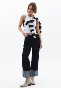 Vit linne, marinblå och krämrandig tröja knuten runt halsen, mörk denim vida byxor med kontrasterande fåll, beige ankelstövlar.