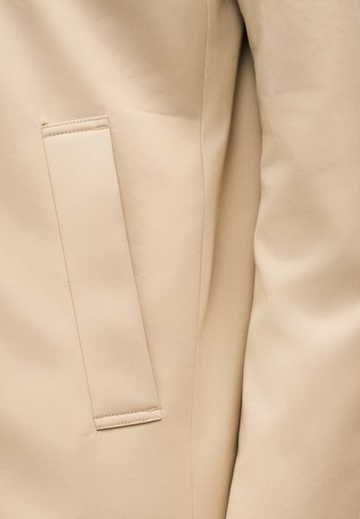 Beige jakkeærme og lodret slidse lomme med syningsdetalje på glat stofoverflade.