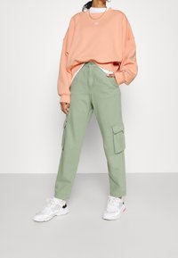 Persikofärgad oversized sweatshirt med vitt logotyp, gröna cargobyxor med sidofickor, och vita sneakers med grå och rosa accenter.
