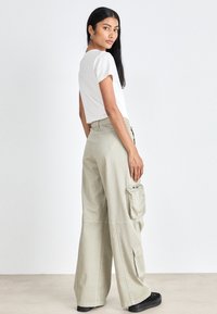 Light khaki cargo pants with wide legs, large side pocket, and front zip detail; paired with a fitted white short-sleeve shirt.