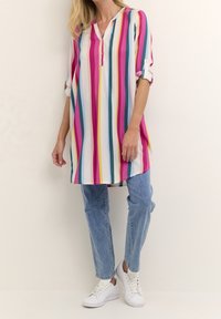 Tunique rayée en rose, bleu, jaune et blanc. Dotée d'une patte de boutonnage, de manches retroussées et d'une coupe ample. Associée à un jean bleu clair et des baskets blanches.