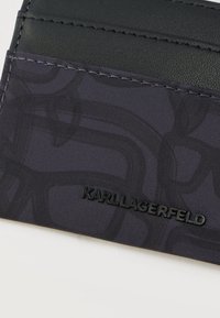 Tuvplāns melnam kartes turētājam ar auduma un ādas daļām, kurās redzami smalki raksti un melns Karl Lagerfeld logotips.