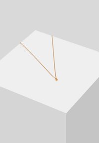 Kuldtooni ahelkaelakee omab õrna disaini koos väikese sfäärilise ripatsiga, mis puhkab valgel geomeetrilisel pinnal.