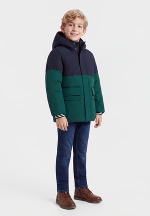 Junge steht und lächelt, trägt eine blau-grüne Kapuzen-Winterjacke, blaue Jeans und braune Lederstiefel.