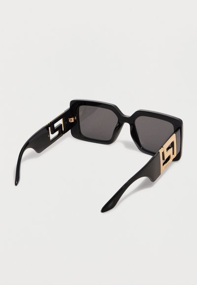 Μεγάλα τετράγωνα μαύρα γυαλιά ηλίου με σκουρόχρωμους φακούς και χρυσά γεωμετρικά διακοσμητικά λογότυπα στα πλαϊνά, εμφανιζόμενα υπό γωνία σε λευκή επιφάνεια.