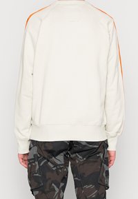 Sudadera beige claro con franjas laterales naranjas, puños y dobladillo acanalados. Vista posterior que muestra textura suave, diseño casual y detalles mínimos.