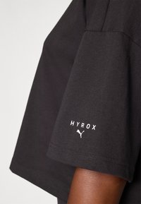 Čierne tričko s krátkym rukávom vyrobené z bavlny, s bielym textom "HYROX" a logom Puma na rukáve, so voľným strihom a hladkou textúrou.
